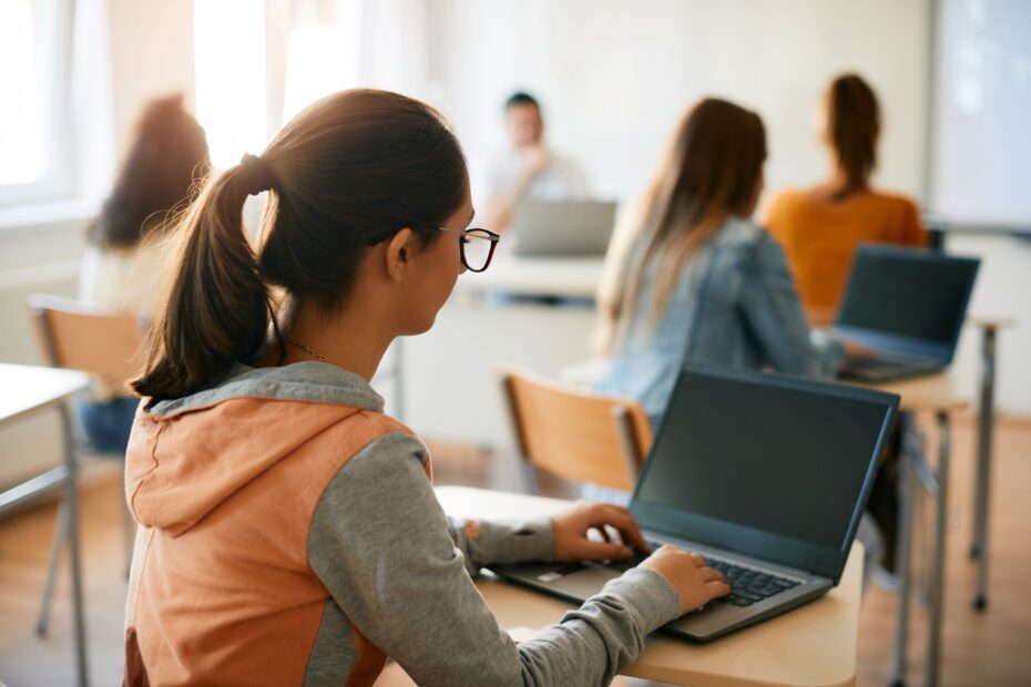 Leerlingen zitten in een klaslokaal en werken op laptops. Op de voorgrond is een meisje met een bril en een staart zichtbaar, gekleed in een grijs-oranje hoodie. Ze typt op haar laptop. Op de achtergrond zitten meer leerlingen aan bureaus, allemaal gericht op hun scherm. De docent zit vooraan in de klas bij een bureau. Het lokaal is licht en modern, met natuurlijk zonlicht dat naar binnen valt.